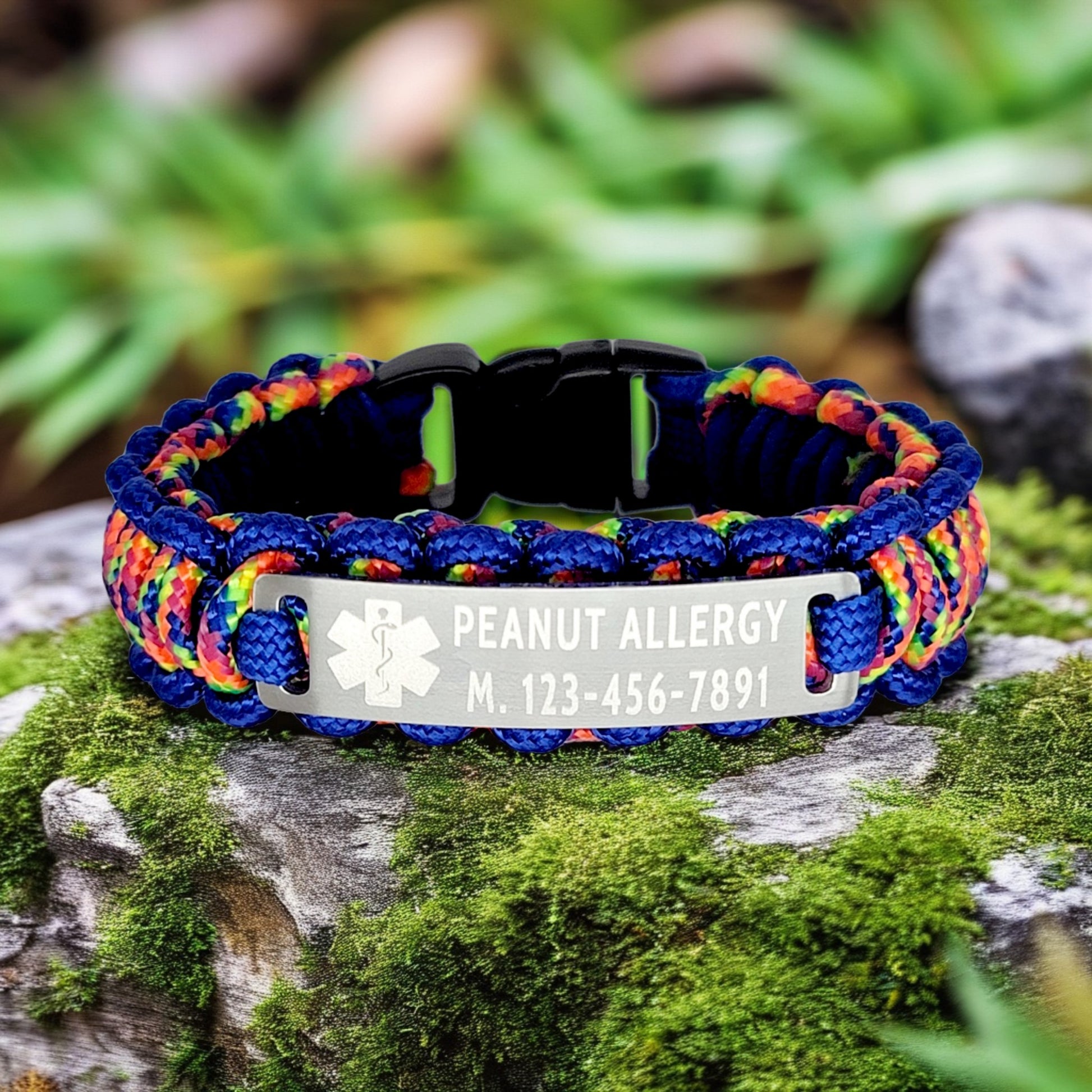 A close-up of a colorful paracord bracelet with a blue and rainbow design. The metal tag is engraved with Peanut Allergy and a medical symbol, resting on a moss-covered rock.