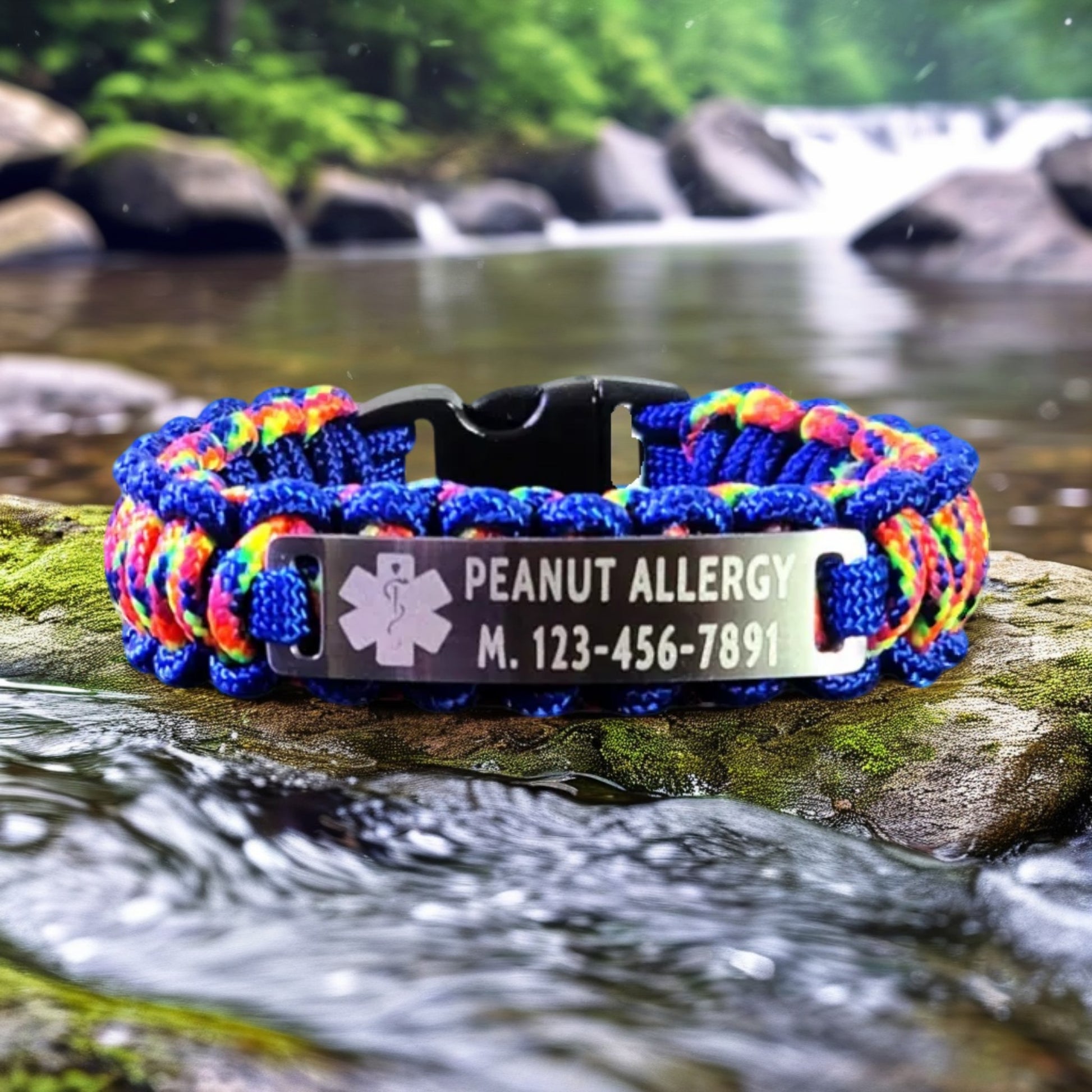 A paracord bracelet with a vibrant blue and rainbow cord design, engraved with Peanut Allergy and a medical symbol, displayed on a rock by a flowing stream.