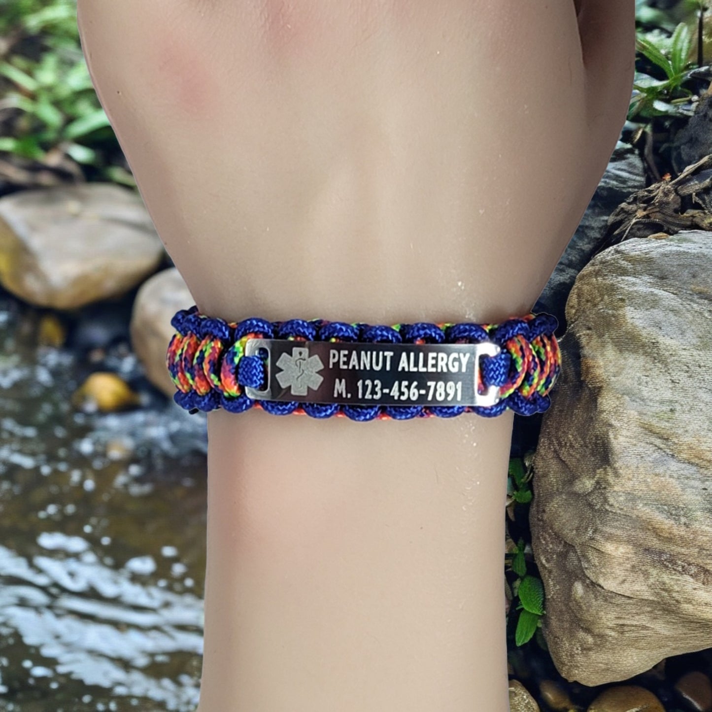 A wrist wearing a paracord bracelet with blue and rainbow cord and a metal tag engraved with Peanut Allergy and a medical symbol, set against a natural rocky background with a stream.